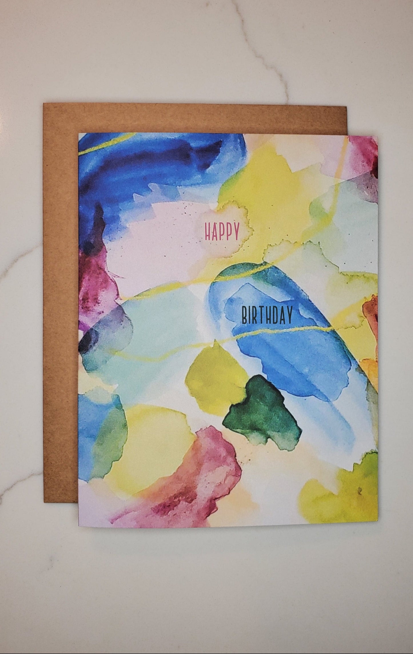 A greeting card with a colorful watercolor design, featuring a variety of bright colors and the text 'HAPPY BIRTHDAY' on the front, presented on a marble surface.