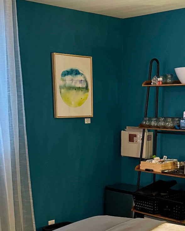 Room interior with teal walls, a treatment room bed, and various items on shelves. Acupuncture studio calm serene treatment room, green planet art on wall.