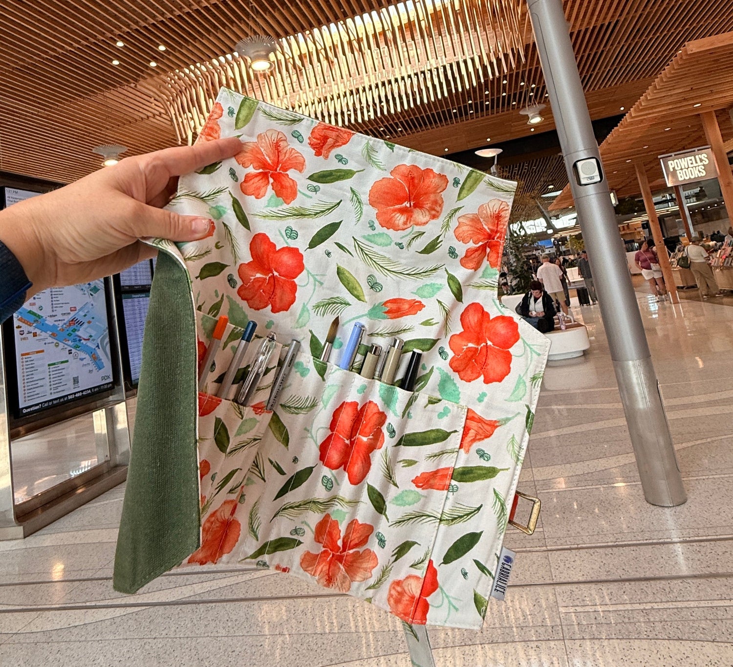 A hand holds a floral travel organizer pouch in a modern indoor airport or transit space with a slatted wood ceiling and tiled floor. The pouch has a white background with red-orange flowers and green leaves, plus multiple pockets and a green fabric loop.