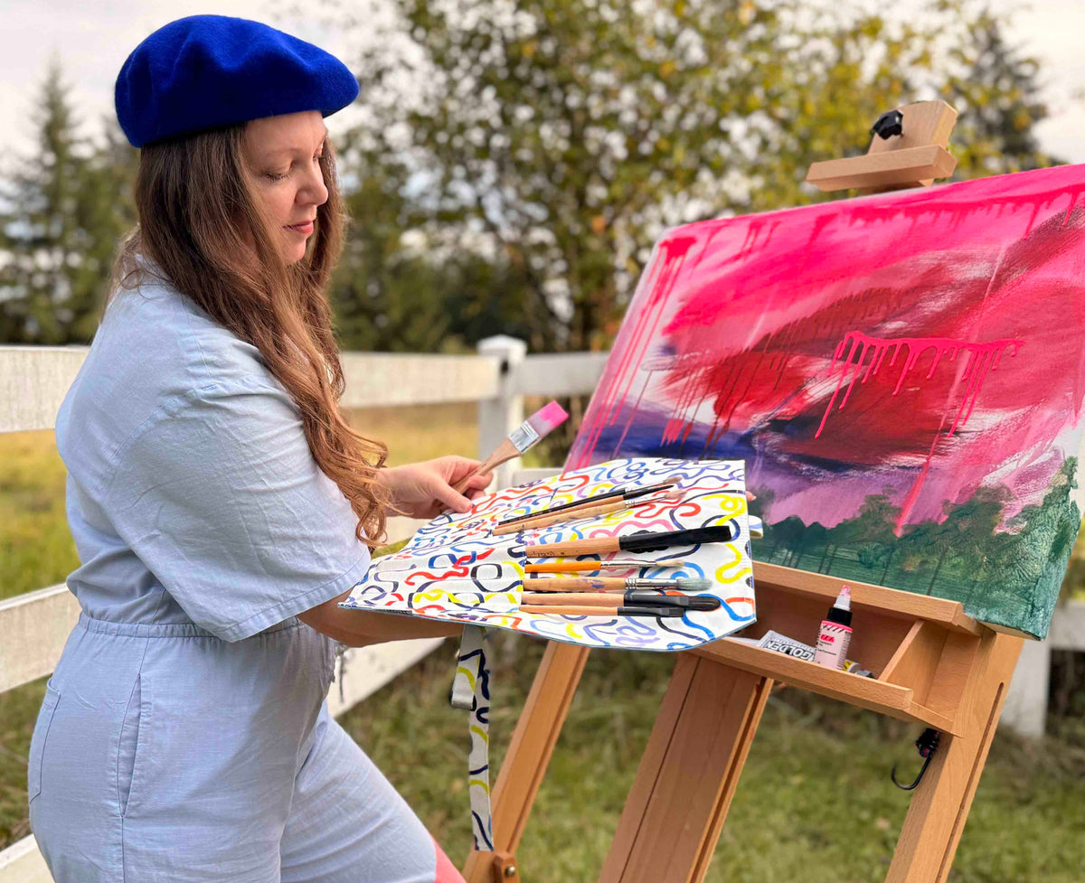A woman wearing a blue beret and light blue outfit stands outdoors beside an easel, painting on canvas. In her hand she's holding a paintbrush roll case with vibrant printed watercolor pattern on the inside. The easel holds a partially painted canvas with vivid pink, red, and green brushstrokes, and a palette with paintbrushes rests on the easel.