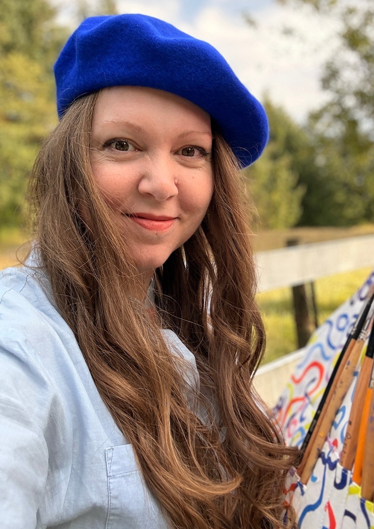 Gabrielle Alice smiles naturally with a deep blue beret and curled hair and a brush roll in her hands with paintbrushes