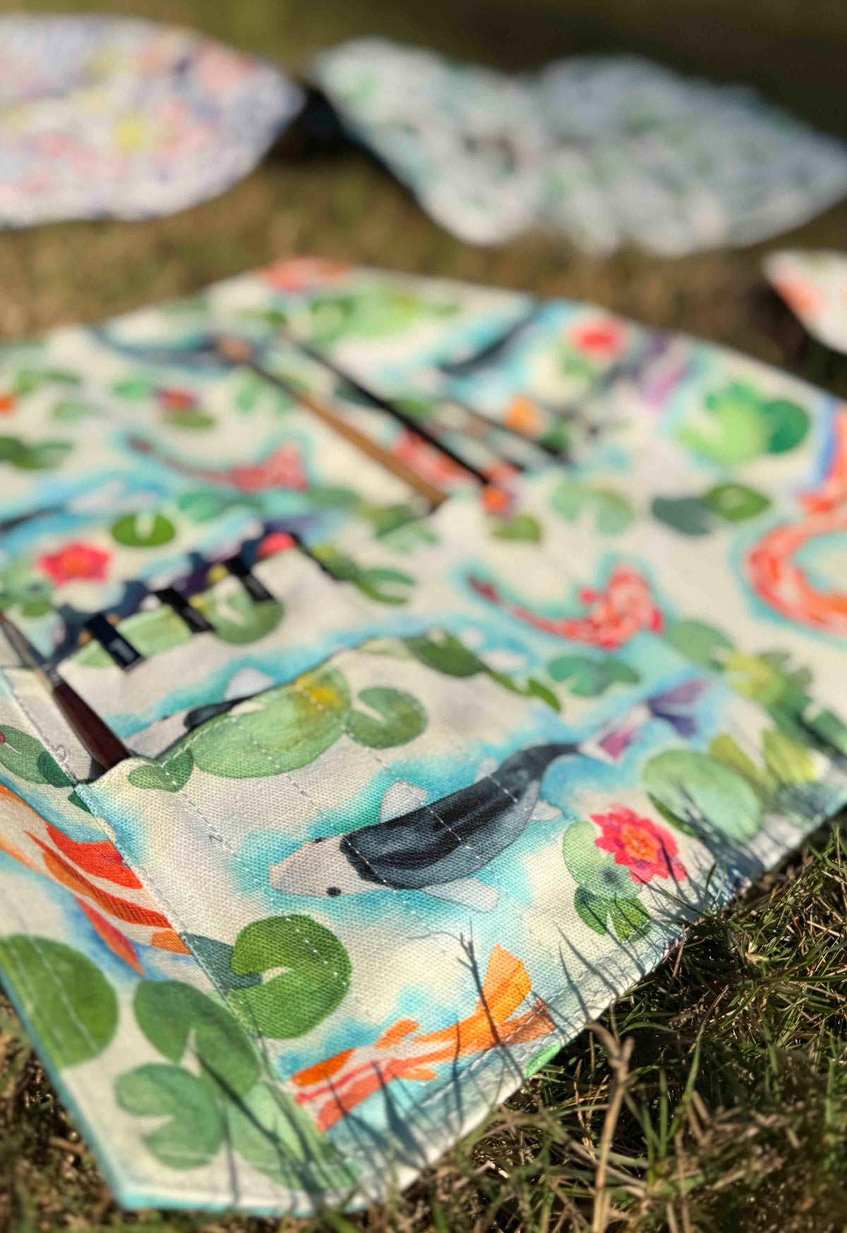 A close-up of a colorful printed fabric pouch or organizer laid on grass, with a watercolor-style design featuring green lily pads, orange fish, blue water, and dark bird silhouettes. Other similar patterned items are blurred in the background.