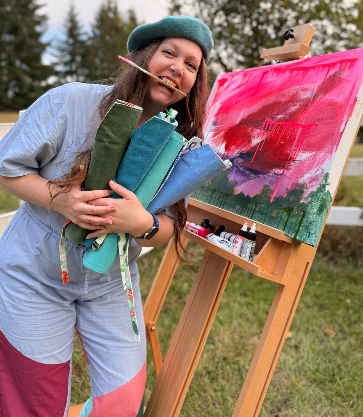 A woman outdoors next to an easel holding a colorful abstract painting in progress. She is wearing a green beret and light blue jumpsuit with pink color-blocked sections, and she is holding several rolled-up paintbrush canvas roll cases. tool rolls plein air organizer artist tool