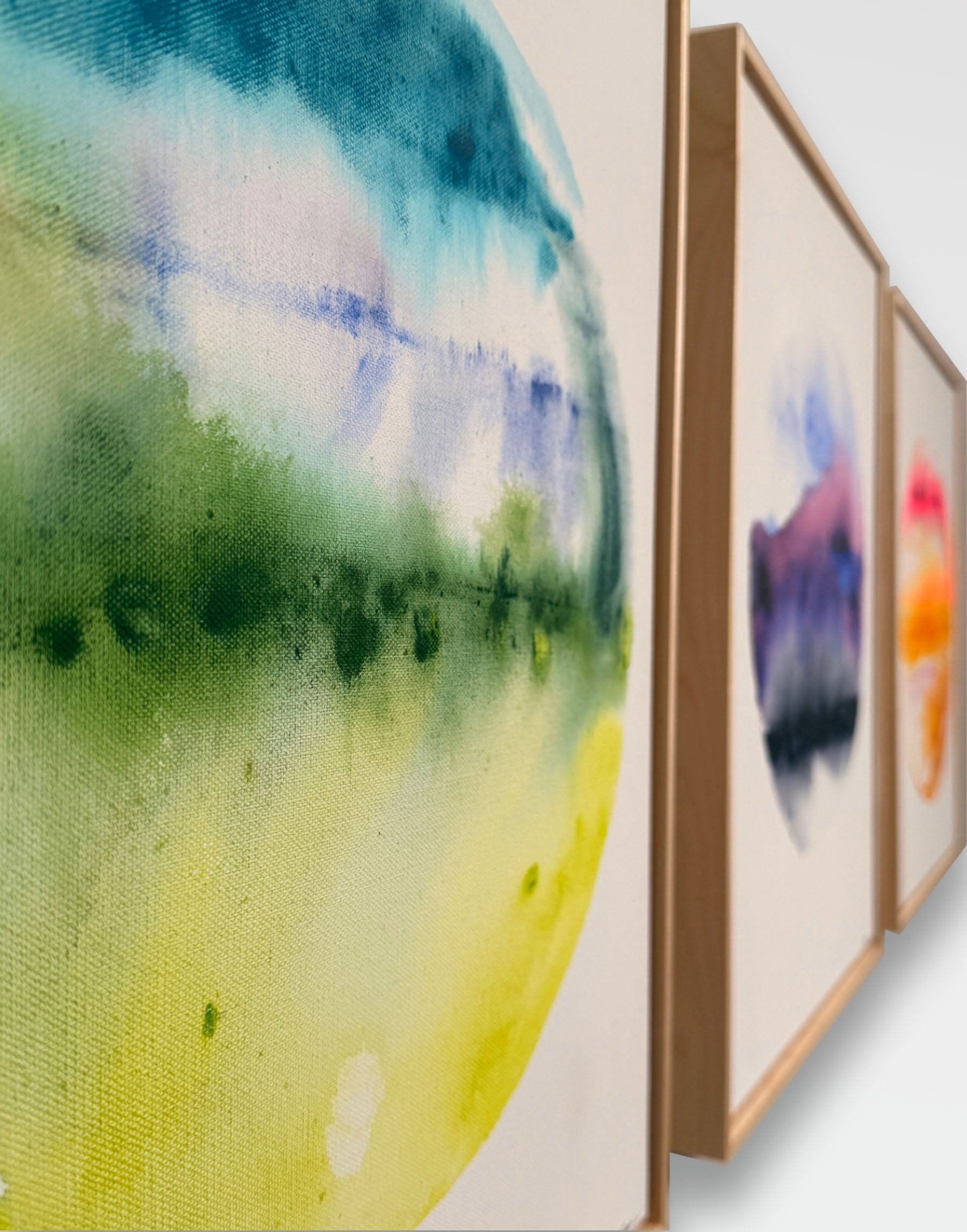 Close up view of maple wood float framed watercolor painting on canvas with circular design in blue, green, and yellow green hues on a white background. with two other pieces in a collection. Purple planet abstract and Orange planet abstract lined up