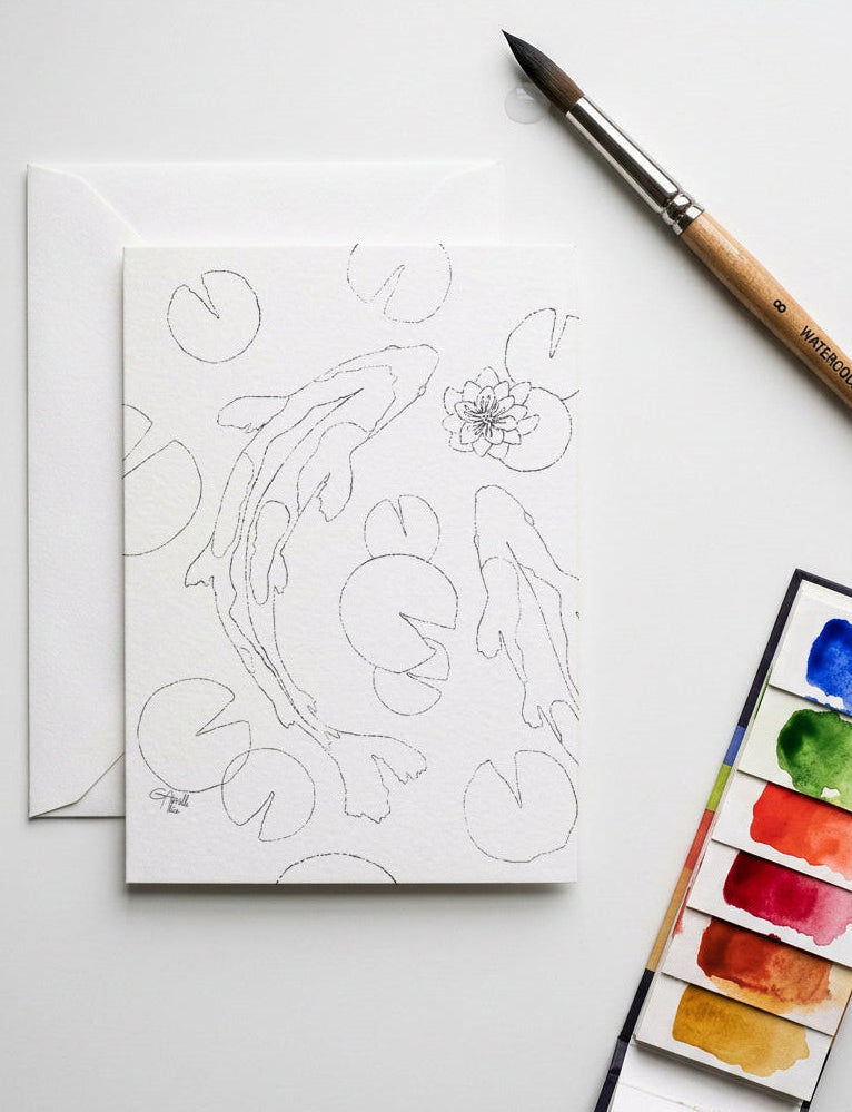 A white art card with black line drawings of koi fish and lily pads, shown with a white envelope on a light gray surface. A paintbrush and a watercolor palette with painted color swatches are placed nearby as styled props.