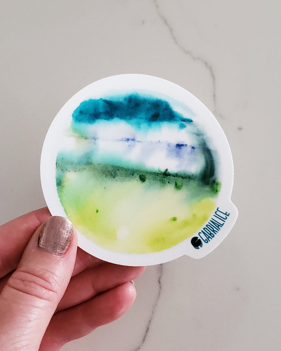 A die-cut sticker with a green and blue watercolor design representing a fantasy planet.