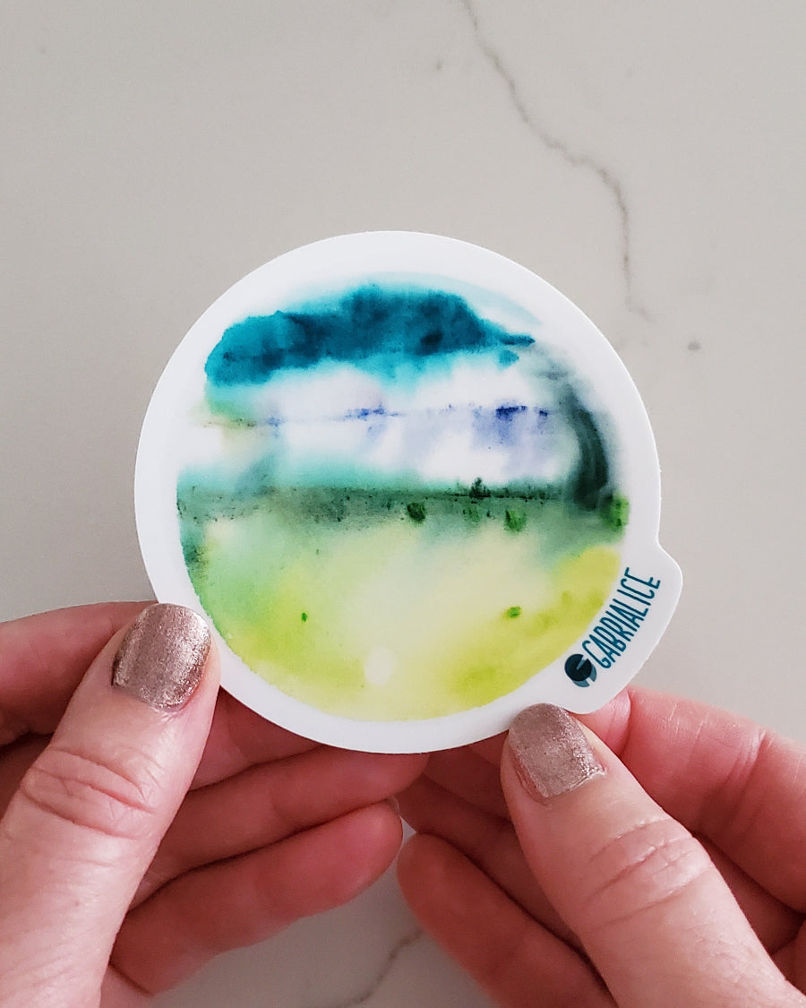 A die-cut sticker with a green and blue watercolor design representing a fantasy planet.
