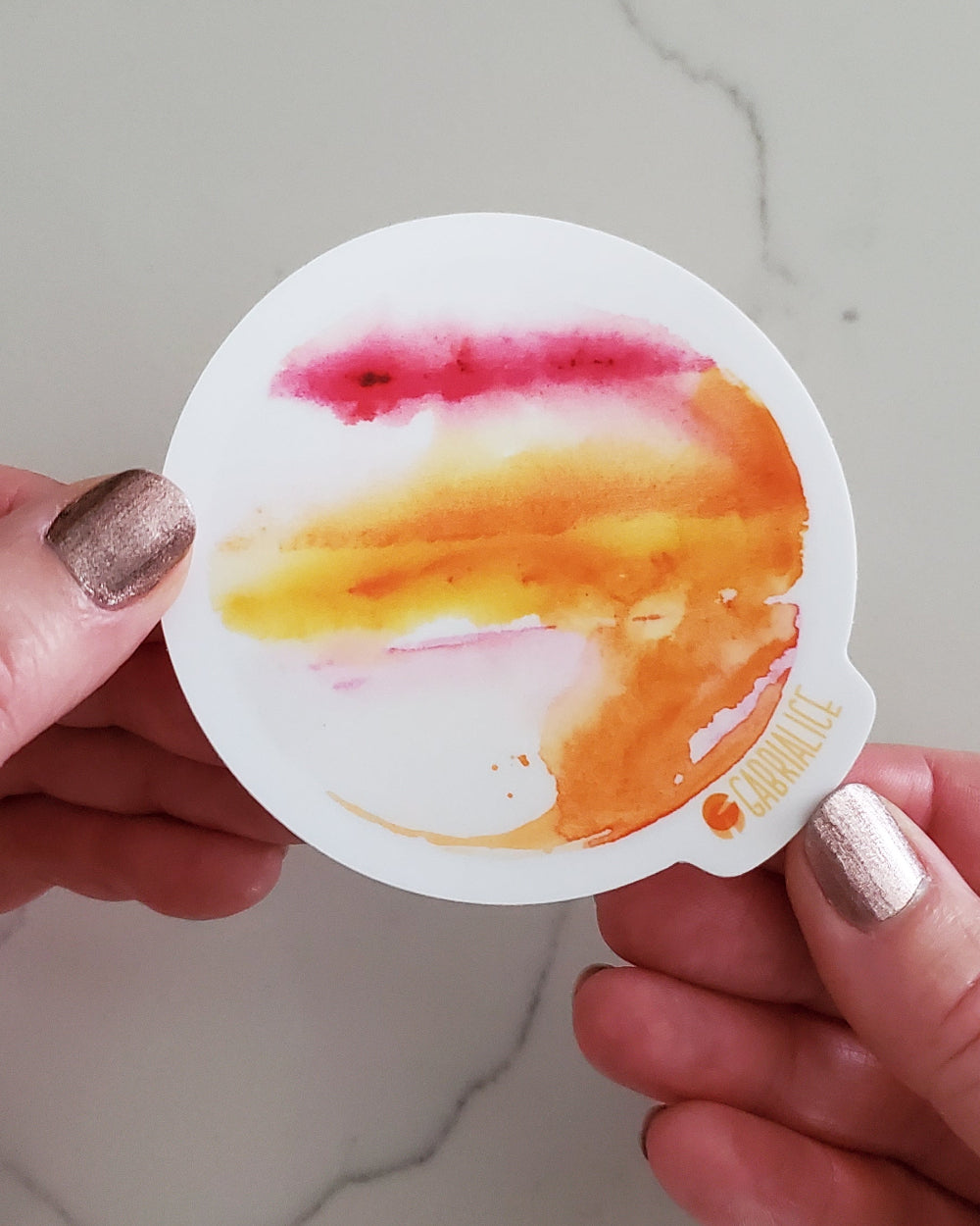 A round vinyl sticker with a colorful design representing a fantasy planet, held between fingers with a marble background.